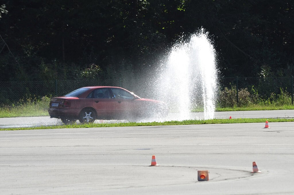 Ein rotes Auto fährt durch Wasserfontänen auf einer Teststrecke, umgeben von grüner Natur.