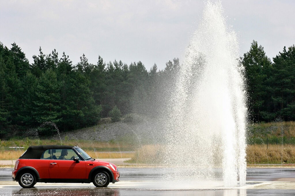 Roter Mini Cooper fährt neben einem hohen Wasserstrahl auf nasser Straße, im Hintergrund Wald.